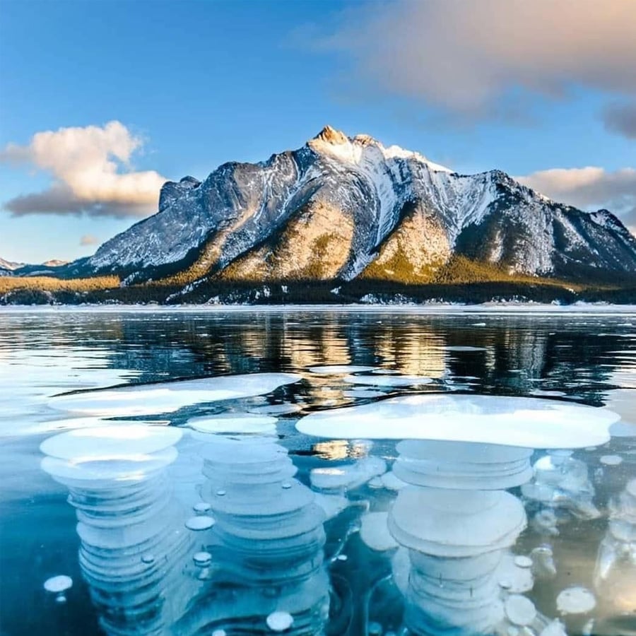 Frozen lake with trapped methane bubbles beneath the ice, reflecting a snow-capped mountain and partly cloudy blue sky in the background.