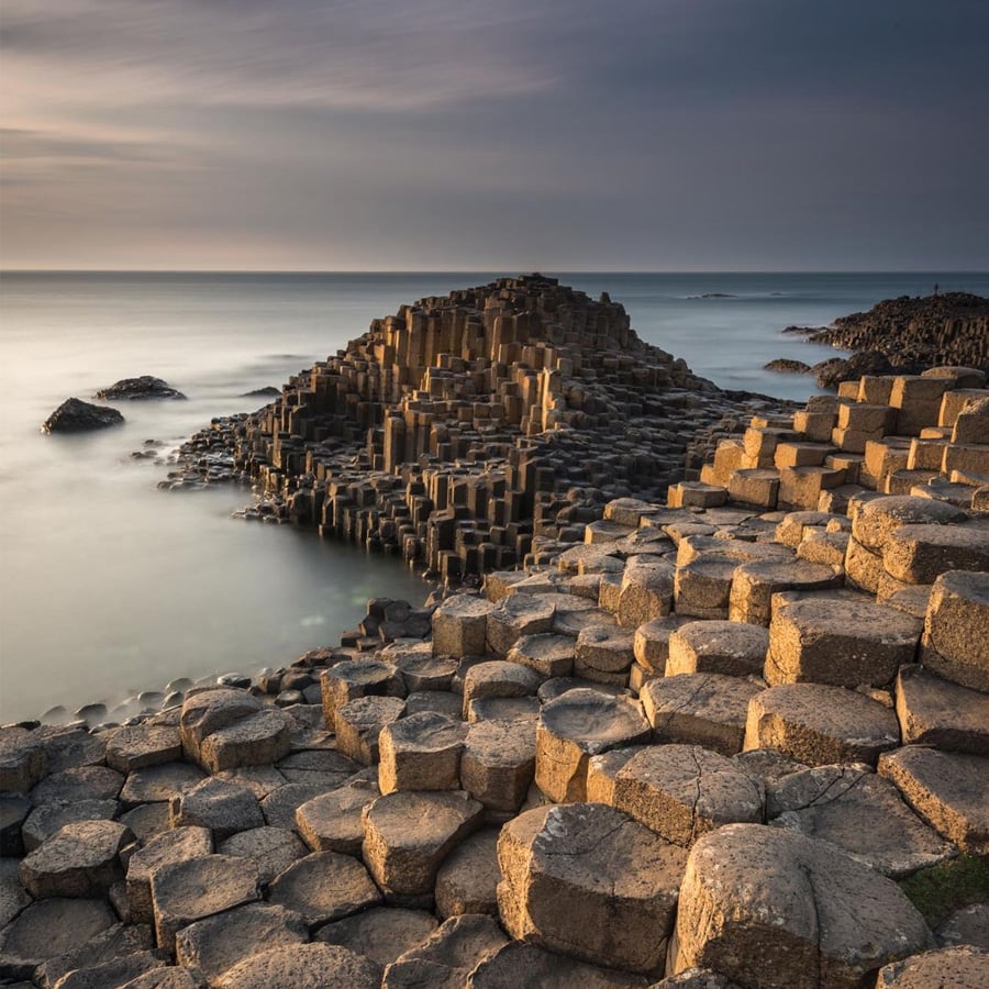 Hexagonal stone columns of the Giant’s Causeway rise from the shore, bathed in golden light. Calm water surrounds the unique rock formations under a cloudy sky.
