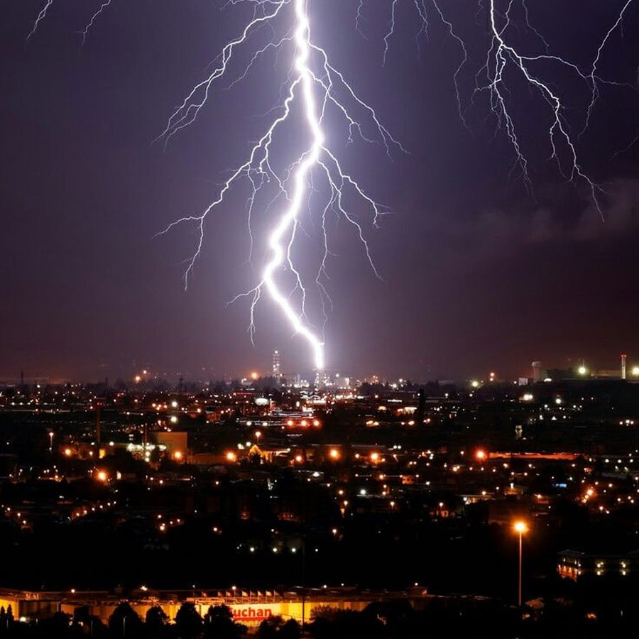 A bright lightning bolt strikes down on a cityscape at night, illuminating the sky and buildings below with branching streaks of light.
