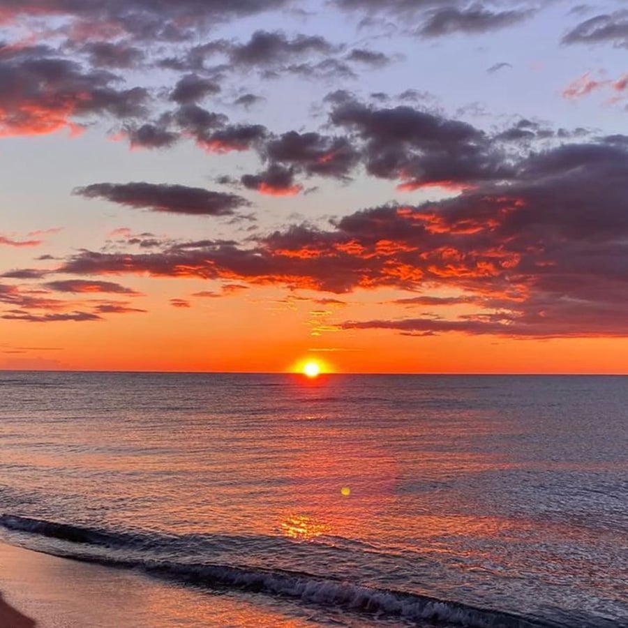 Bright orange sun setting over a calm ocean, with dramatic clouds in a colorful sky of blue, purple, and orange hues. Gentle waves approach the shore in the foreground.