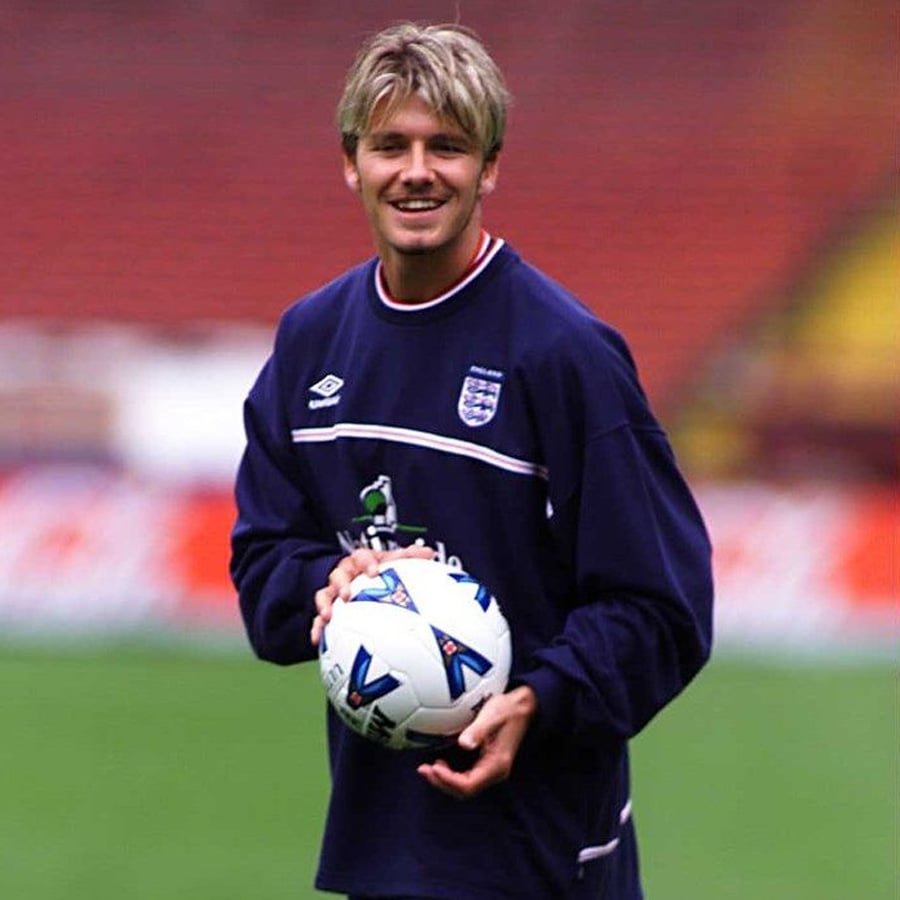 A young man in an England soccer jersey smiles while holding a soccer ball on a field, with stadium seats blurred in the background.