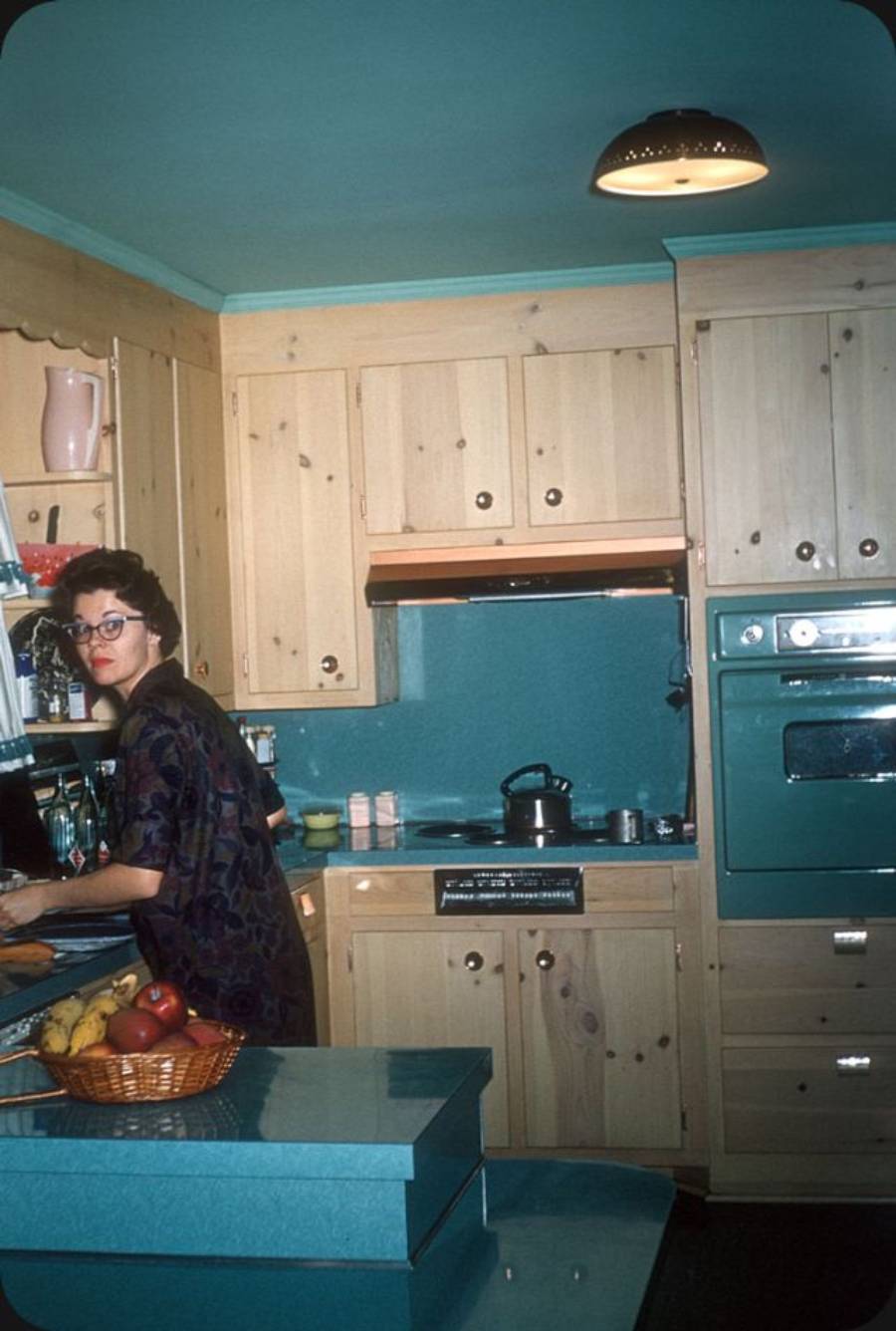 A woman stands in a retro kitchen with wooden cabinets, teal walls, and appliances. She is looking over her shoulder while preparing food. A fruit bowl sits on the counter in the foreground.