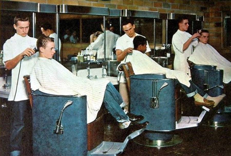 Three barbers in white uniforms cut men's hair in vintage barber chairs. The scene takes place in a brightly lit barbershop with mirrors and sinks in the background.