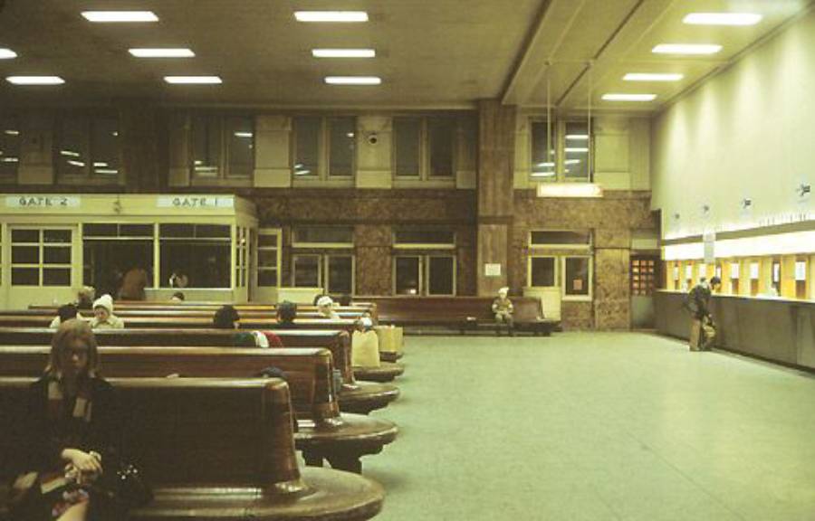 People sit on wooden benches in a spacious, dimly lit waiting area with ticket counters on the right and large windows along the back wall. Some people are waiting, while one stands at a counter.