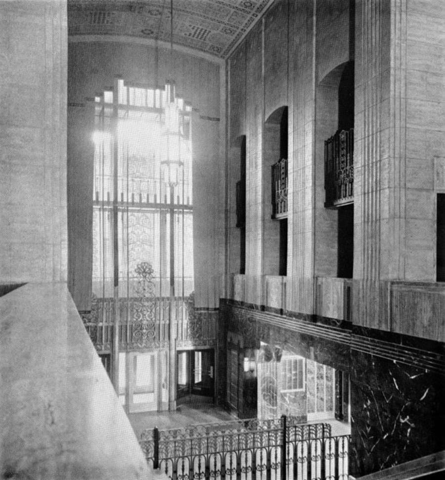 Black and white photo of an Art Deco building interior with tall windows, ornate metalwork, high ceilings, marble walls, and decorative balcony railings. Sunlight streams through the large glass doors and windows.