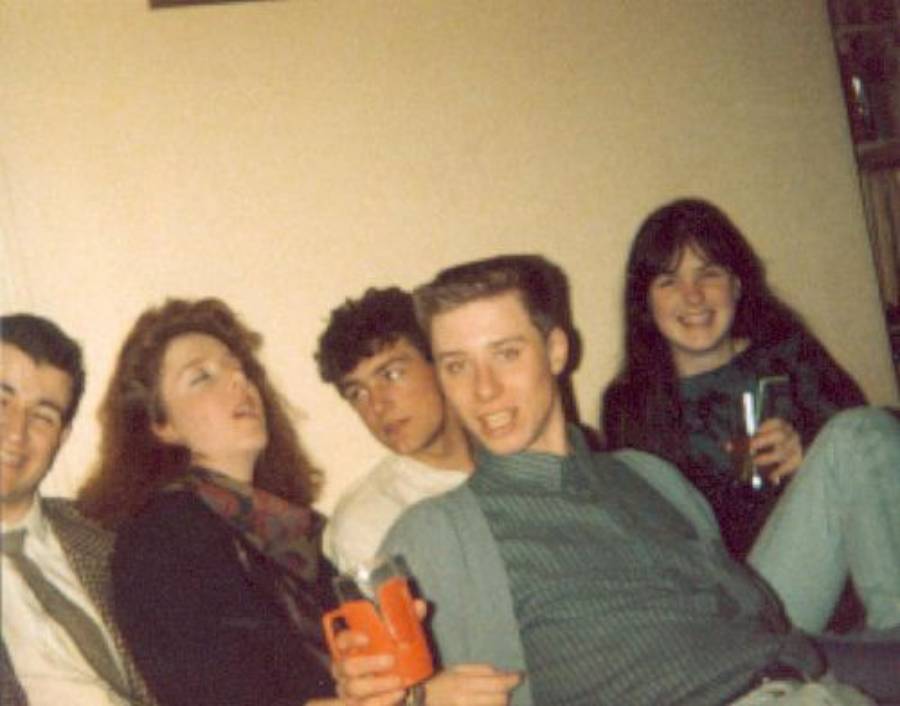 Five young adults sit closely together on a couch, smiling and holding drinks. They appear to be at a casual indoor gathering, with a beige wall behind them and relaxed, cheerful expressions.
