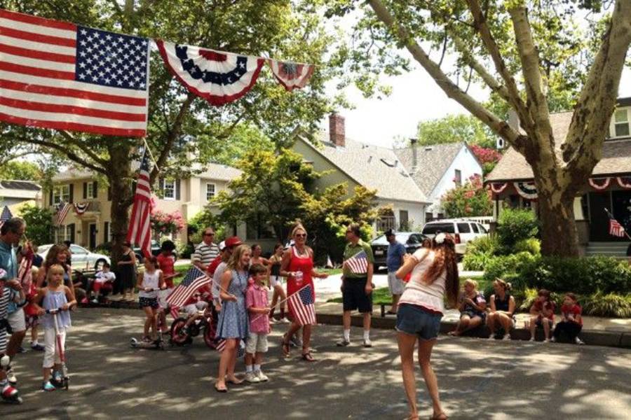 People gather on a sunny street decorated with American flags and bunting, celebrating. Children and adults hold flags, with houses and trees in the background. A woman in front takes a photo.