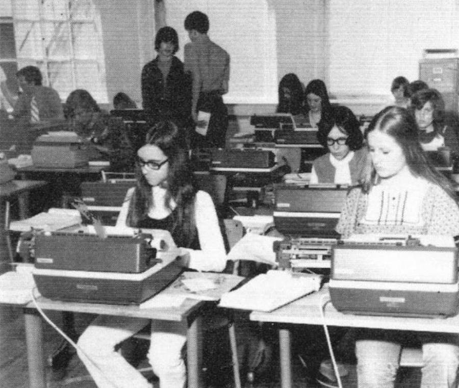 Black-and-white photo of students in a classroom, each working at a typewriter. Some students are typing while others are looking at papers. Shelves and windows are visible in the background.