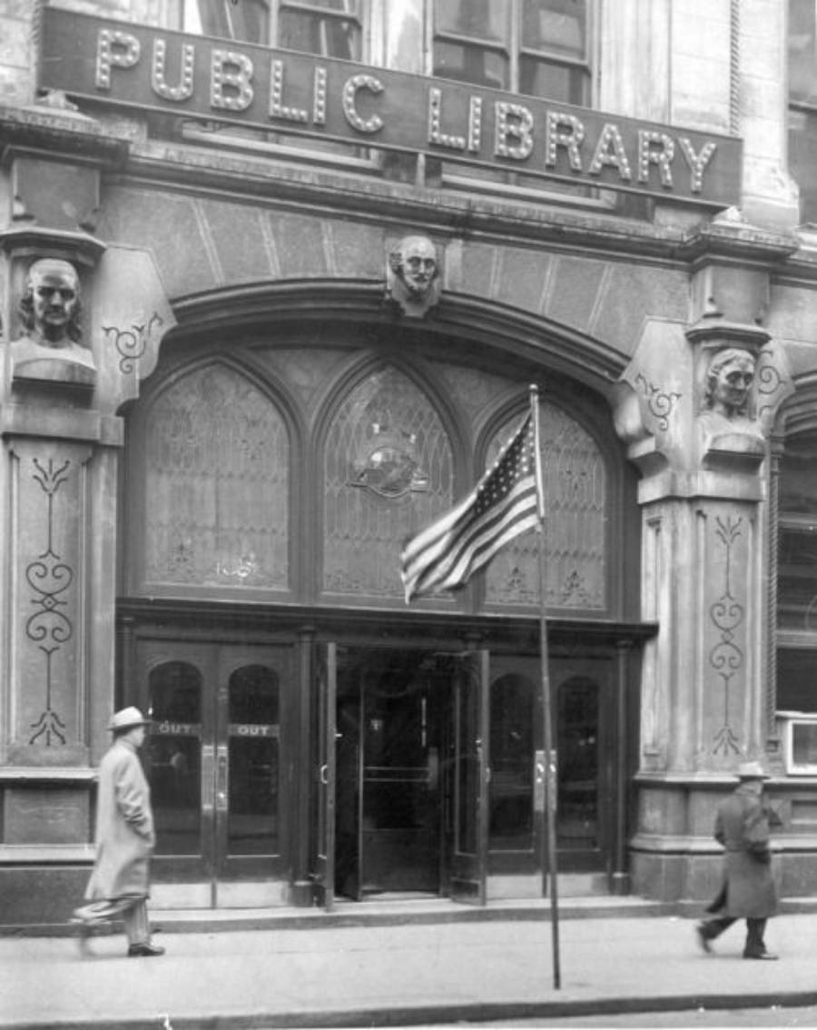 Two people walk past the entrance of a public library, marked by a large sign above ornate doors. An American flag stands on the sidewalk in front of the building.