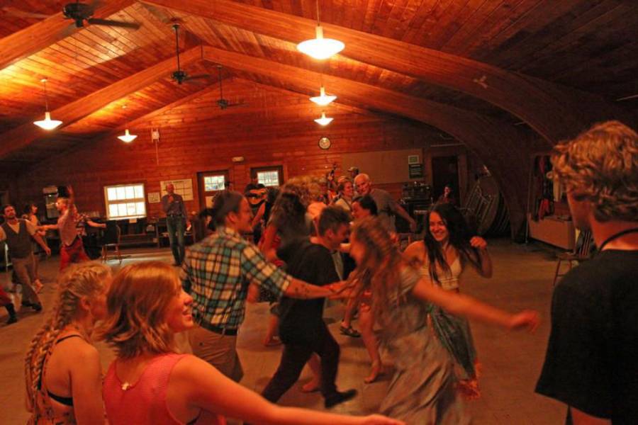 A group of people, some holding hands, dance energetically in a warmly lit wooden room with a high ceiling. The atmosphere appears lively and joyful.