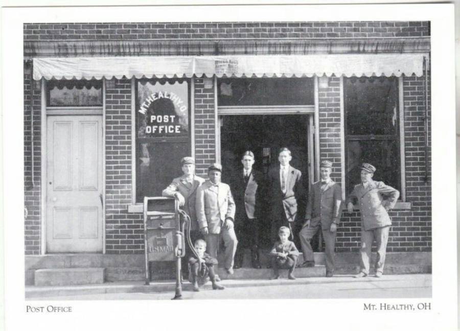 Black and white photo of eight people, including children, standing in front of a brick building labeled "Mt. Healthy Post Office" in Mt. Healthy, Ohio. There is a vintage mailbox in front.