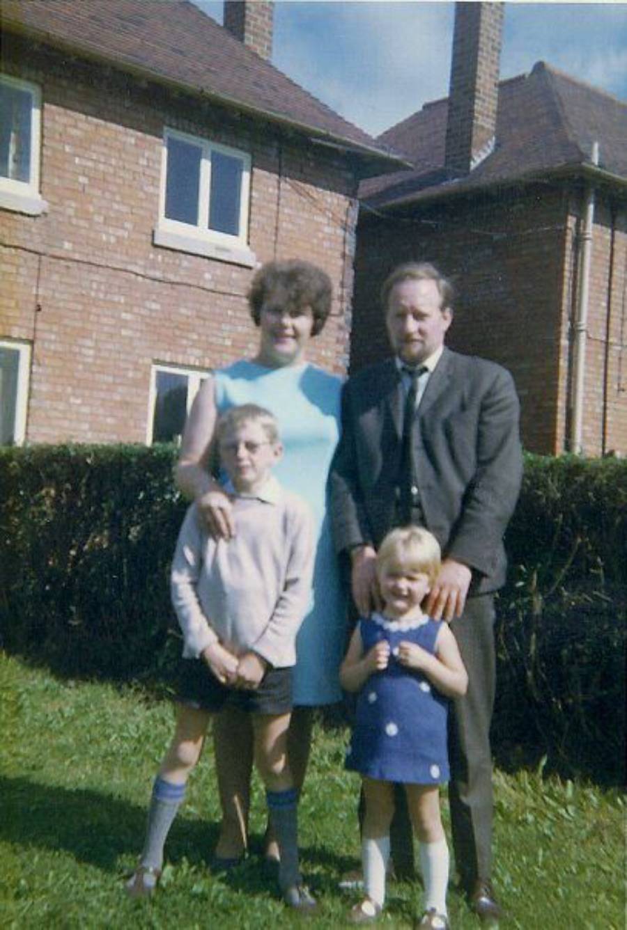 A family of four—two adults and two children—stands on grass in front of a brick house with a hedge. The adults stand behind the children, who are dressed in casual clothes. It’s a sunny day.