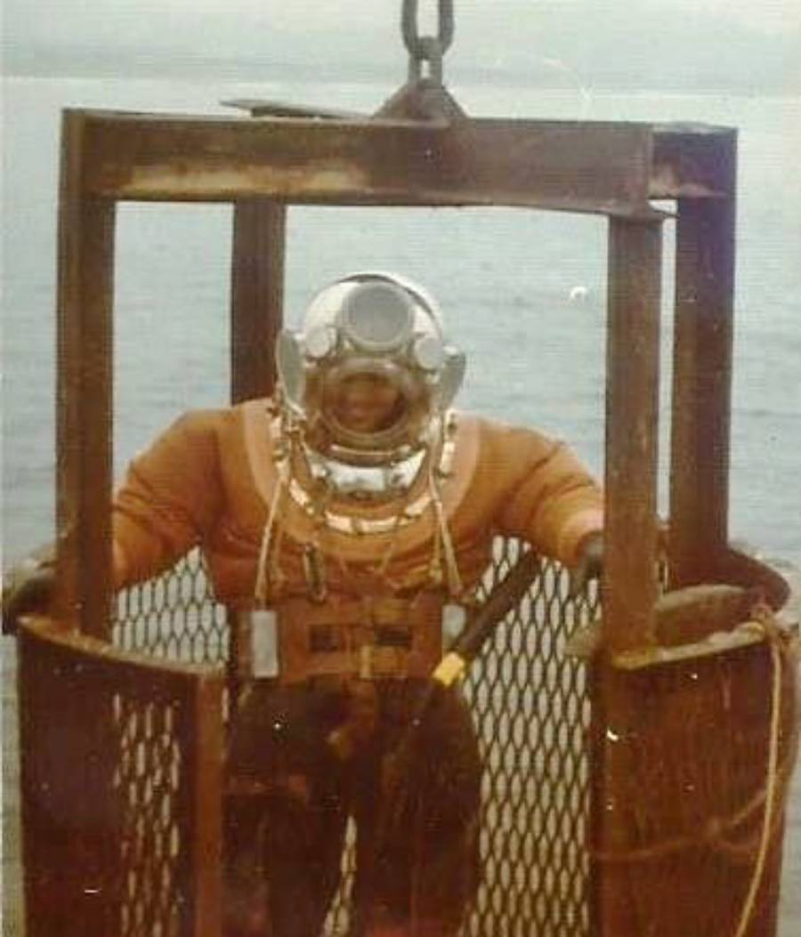 A diver in an old-fashioned, heavy diving suit and helmet stands inside a metal cage suspended above the water, preparing for descent.