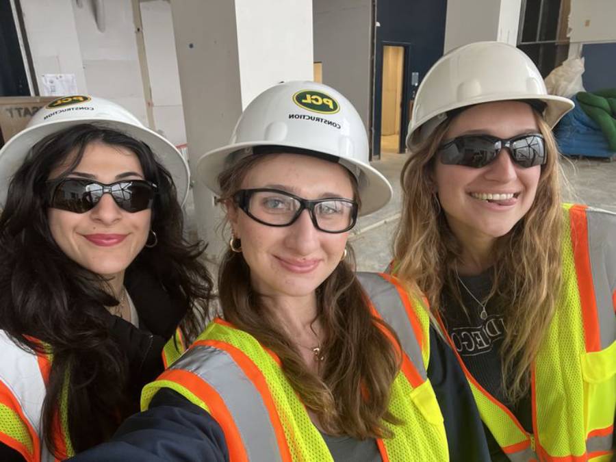 Three women wearing hard hats, safety vests, and protective eyewear smile for a selfie at a construction site.