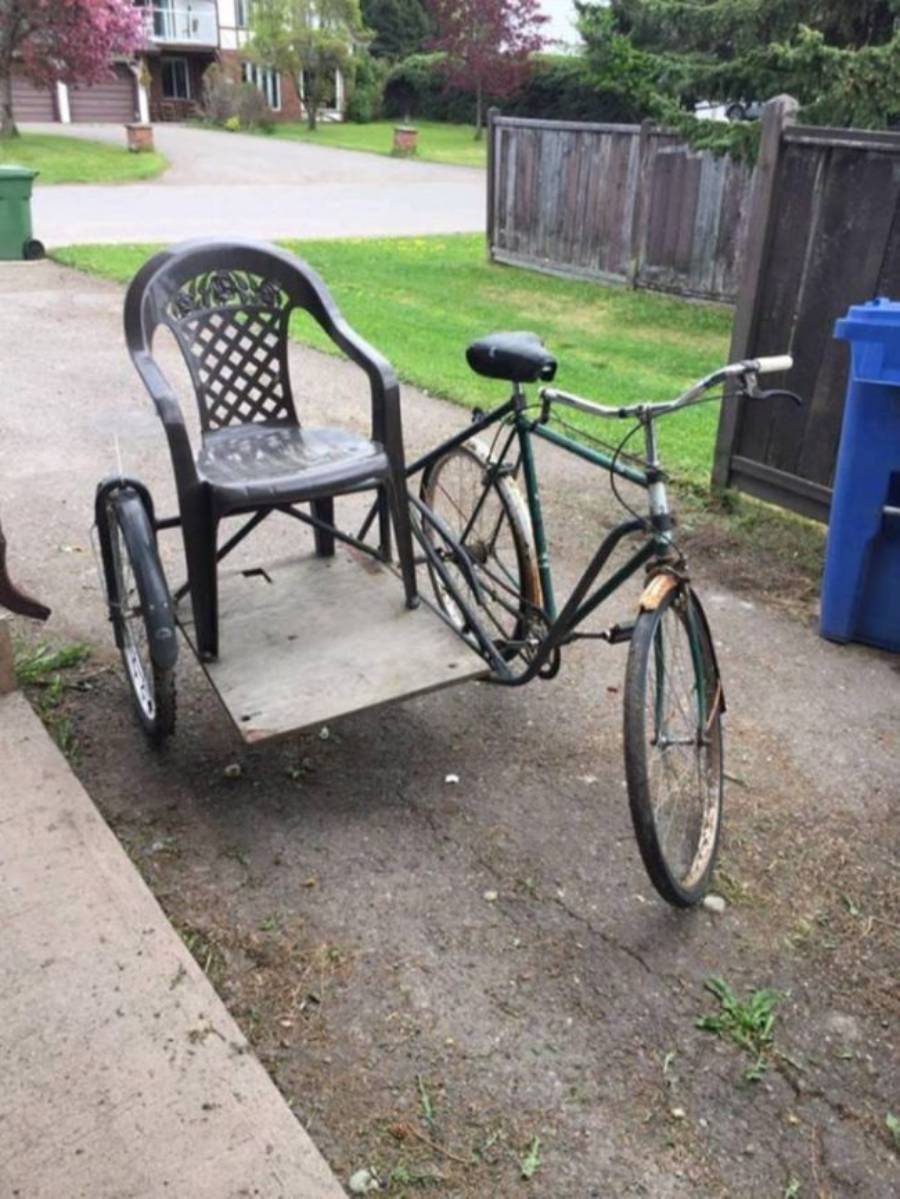 A bicycle modified with a large wooden platform and a plastic patio chair attached to its side, creating a homemade sidecar tricycle, parked on a driveway.