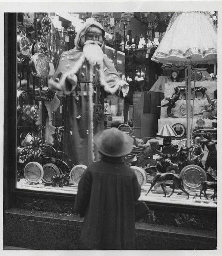 A child in a coat and hat gazes at a festive store window display featuring a large Santa Claus figure, toys, and clocks, evoking a nostalgic holiday atmosphere.