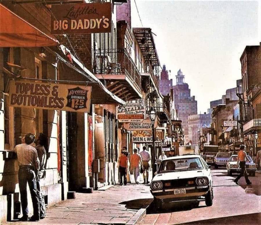 A vintage street scene shows people standing outside clubs and bars with neon signs, including “Big Daddy’s” and “Topless & Bottomless.” A classic car is parked along the sidewalk; multi-story buildings line both sides.