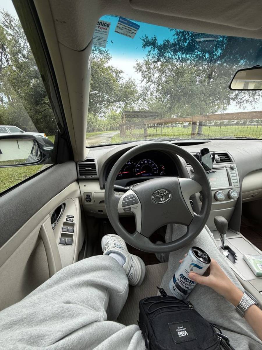 Person sitting in the driver's seat of a Toyota car with legs up, holding a can of C4 energy drink. The car is parked near a wooden fence with trees visible outside. A black bag rests on their lap.