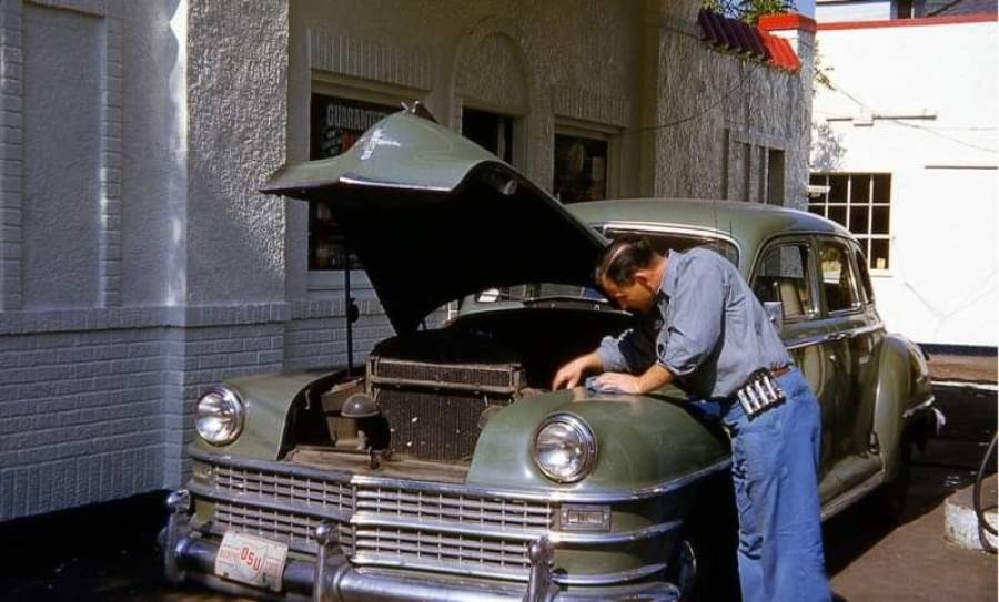 A man in a blue uniform inspects the engine of a vintage green car with its hood open, parked outside a white brick building.
