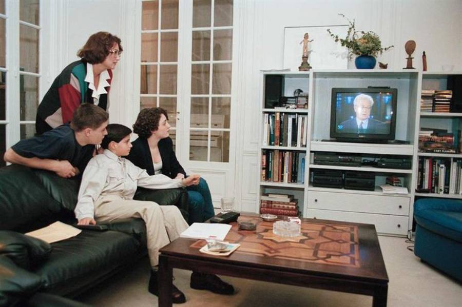 Four people sit in a living room, watching a man speaking on TV. The room has bookshelves, a coffee table, and various decorations. The group looks focused on the broadcast.