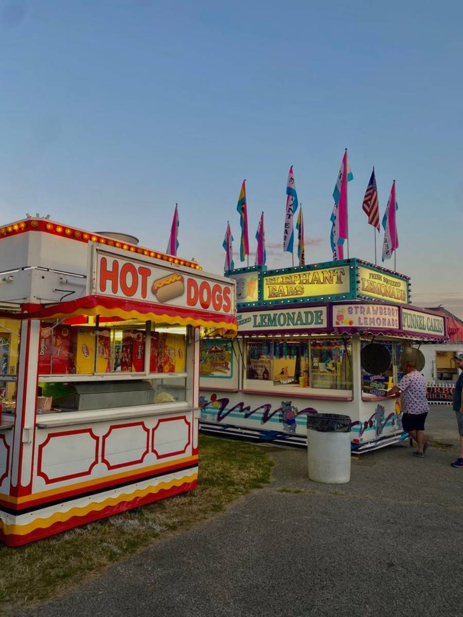 A colorful fairground scene with a hot dog stand in the foreground and a vibrant concession booth selling lemonade, funnel cakes, and elephant ears in the background. Flags are displayed on top; a person stands nearby.
