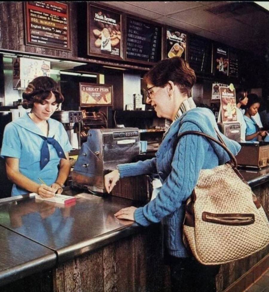A woman in a blue sweater places an order at a retro fast-food counter, while a cashier in a blue uniform writes on a pad. Menus and another worker are visible in the background.