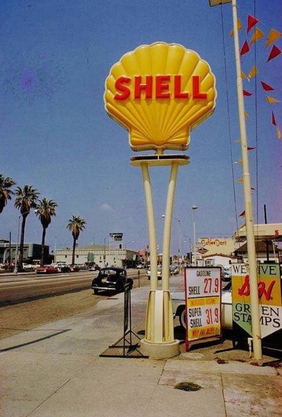 A vintage Shell gas station sign shaped like a yellow shell stands by a road lined with palm trees, with old cars and a price board showing gas prices below.