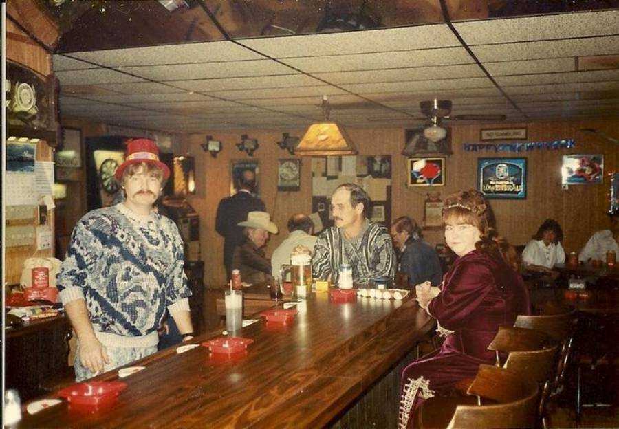 A man in a red hat stands behind a bar. A man and woman sit at the bar facing the camera. Several other people sit at tables in the background. The bar has wood decor and vintage lighting.