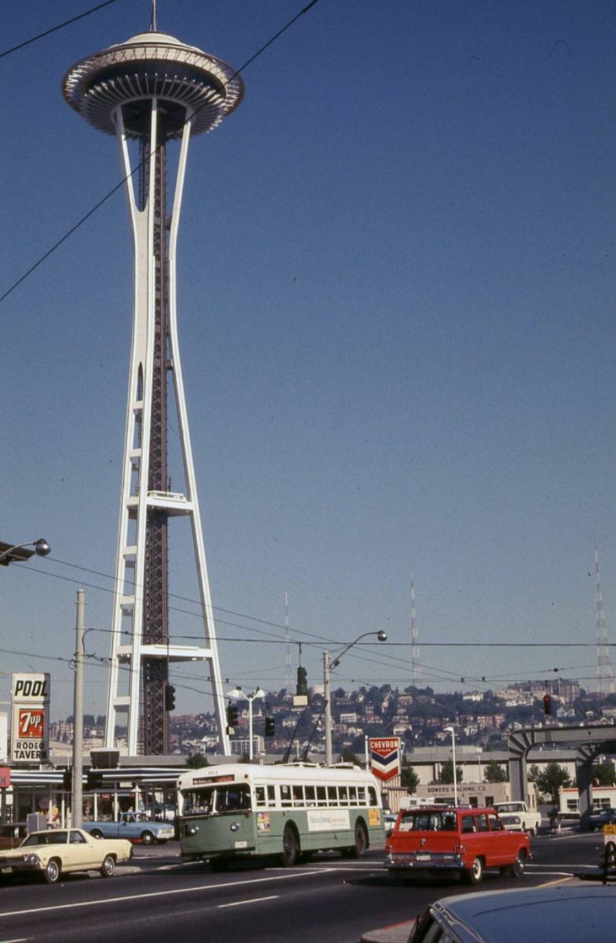 The photo shows the Seattle Space Needle against a clear blue sky, with cars and a green-and-white bus driving on a busy street below. A 7UP sign, gas station, and various power lines are also visible.