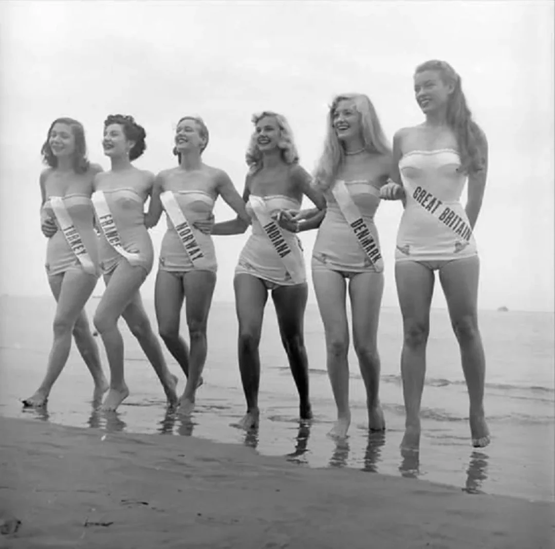 Six women in swimsuits and sashes labeled with country names stand smiling in a row on a beach, ankle-deep in water, holding each other’s waists. The photo is black and white, suggesting it is from a past era.