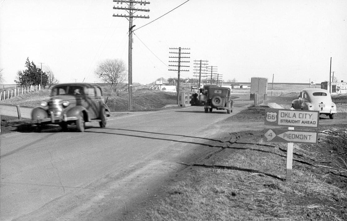 Black and white photo of 1930s cars driving on a rural road near a Route 66 sign pointing to "OKLA CITY" and "PIEDMONT," with power lines and fields in the background.