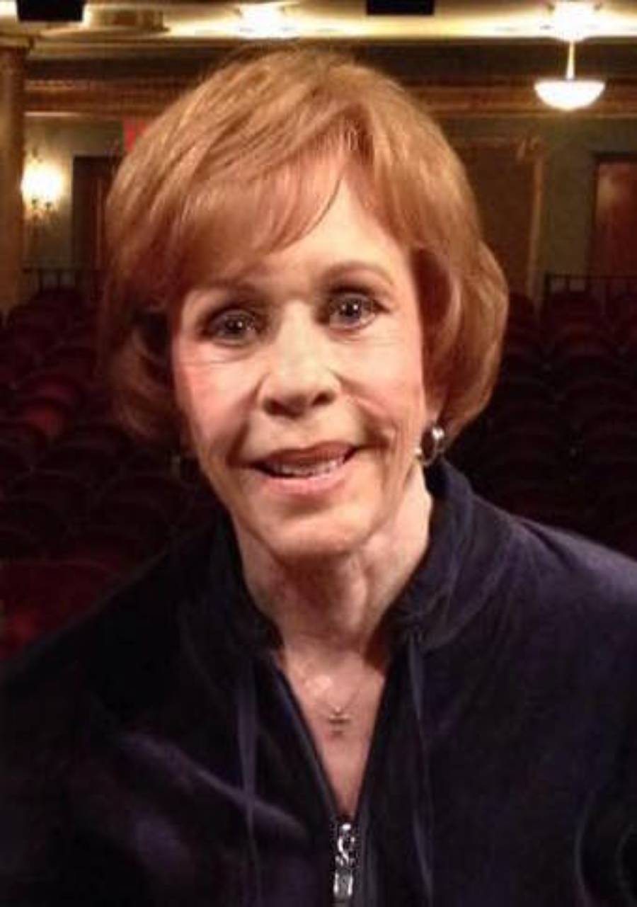 A woman with short, reddish-brown hair and a warm smile stands indoors, wearing a dark top and earrings. The background shows empty theater seats and soft lighting.