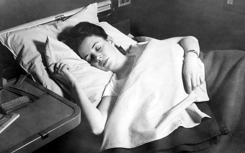 A woman lies asleep on her side in bed, covered with a blanket up to her chest, holding a pillow. A table with a hairbrush and other items is beside her. The photo is in black and white.