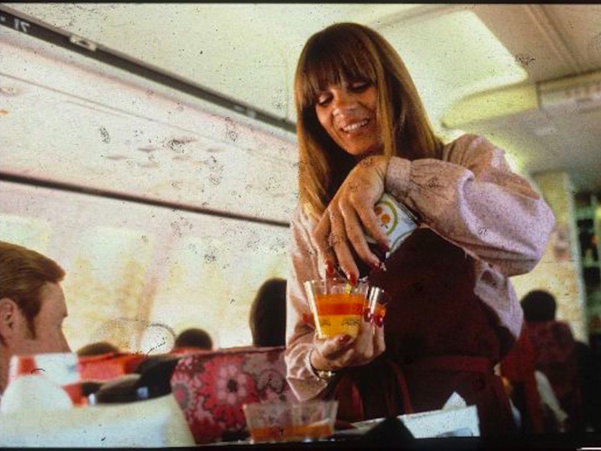 A flight attendant with long hair smiles while pouring a drink into a cup for a passenger on an airplane. The cabin has red-patterned seats and windows letting in daylight.