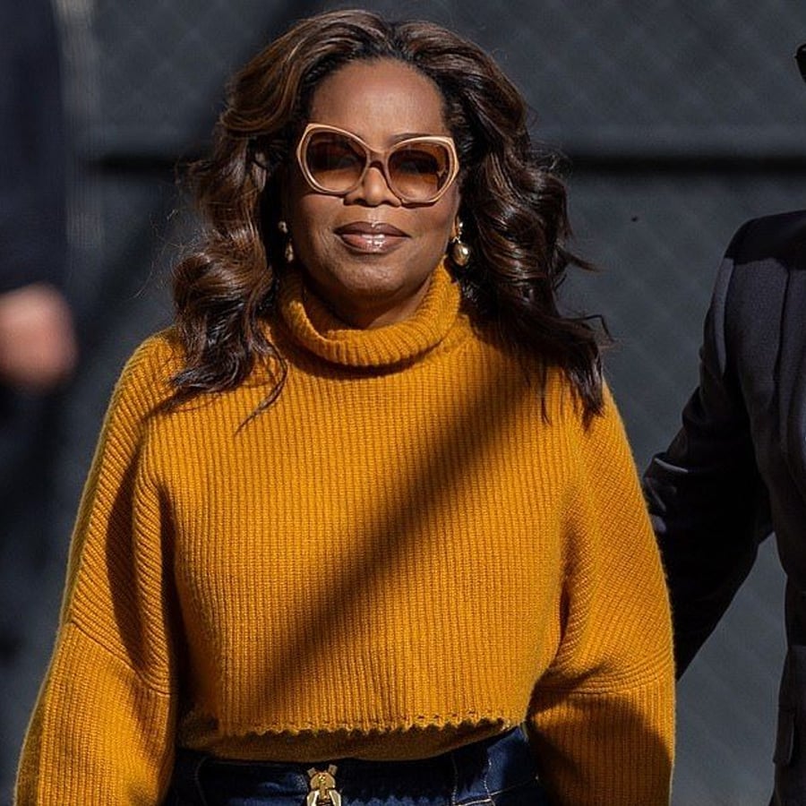 A woman wearing large sunglasses and a mustard yellow turtleneck sweater stands outdoors, smiling slightly. She has long, wavy hair and is accessorized with earrings.