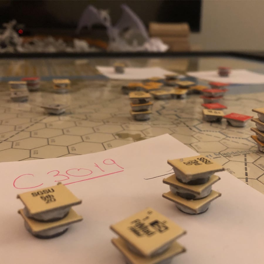 Hex grid game board with stacked game pieces, some labeled "SODU 911" and "HQ," on top of a paper with "C3019" written in pink. Other game pieces and a dragon miniature are in the background.
