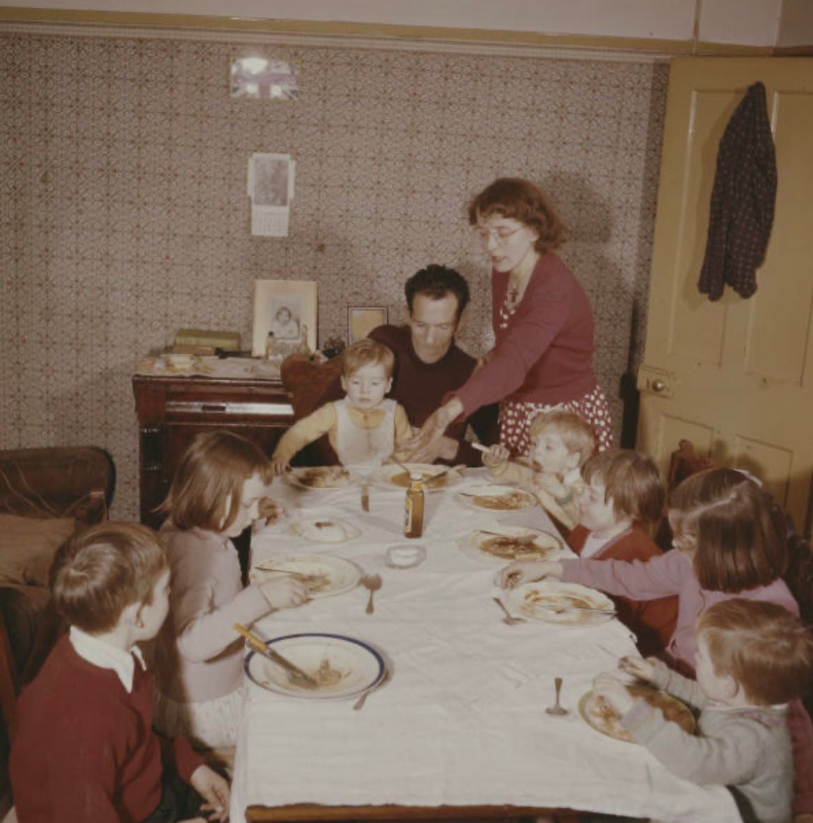 A family of nine, including several children, gathers around a dining table set with food in a cozy, vintage-style room. An adult serves food while everyone prepares to eat.