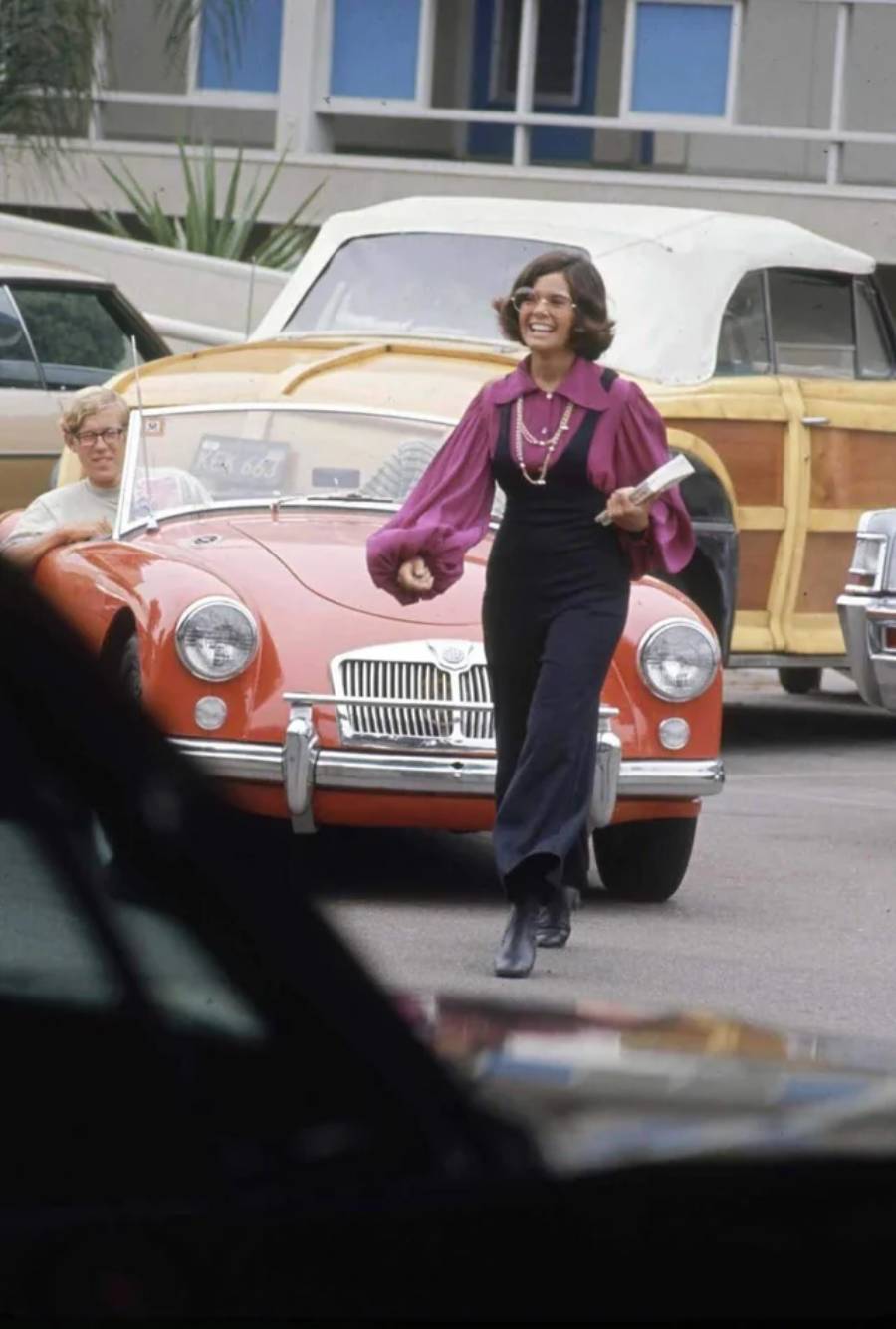 A woman in a purple blouse and dark overalls walks and smiles while holding books in front of a vintage red convertible. Another person sits in the car. Classic cars and a building are visible in the background.