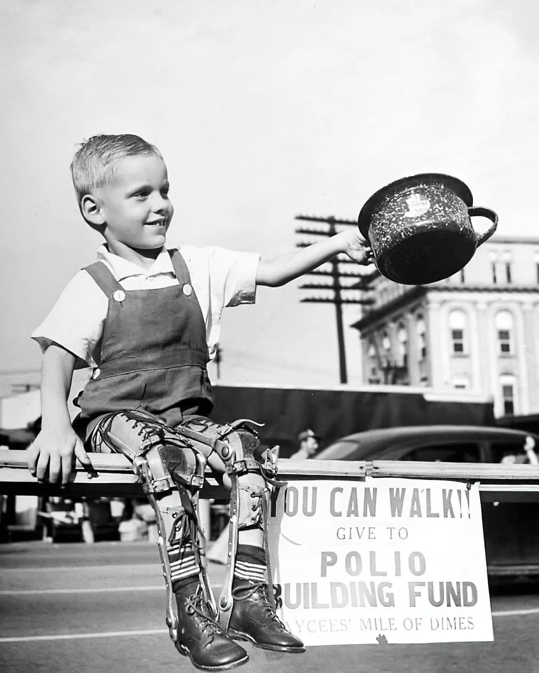 A smiling young boy with leg braces sits on a railing, holding a collection pot. A sign below reads, "You can walk!! Give to polio building fund." Cars and buildings are visible in the background.