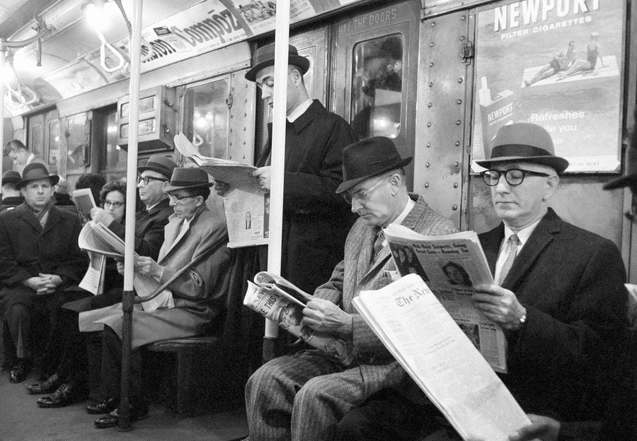 Black and white photo of men in suits and hats sitting and standing in a subway car, reading newspapers. The train walls have old ads, including one for Newport cigarettes.