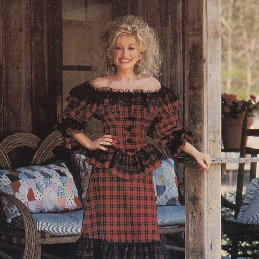 A woman with curly blonde hair, wearing a red and black plaid off-shoulder dress, stands smiling on a rustic porch beside wicker furniture with patchwork cushions and potted flowers in the background.