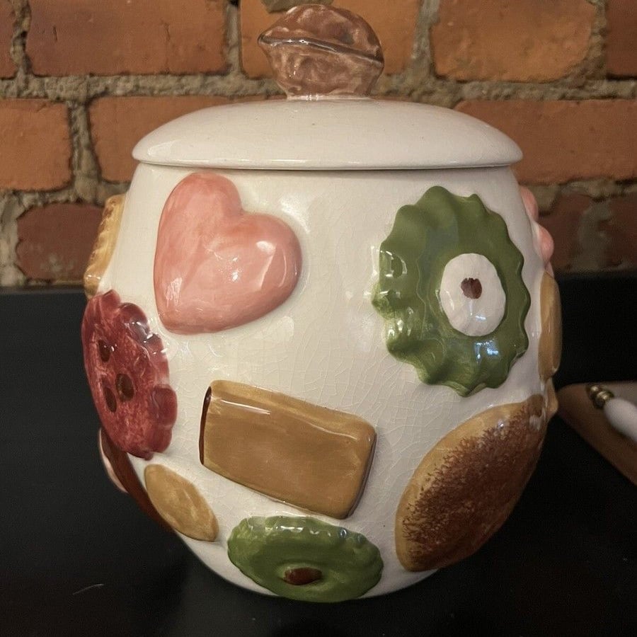 A ceramic jar with a lid, decorated with colorful, raised shapes resembling cookies, hearts, and circles, sits in front of a brick wall on a dark surface.