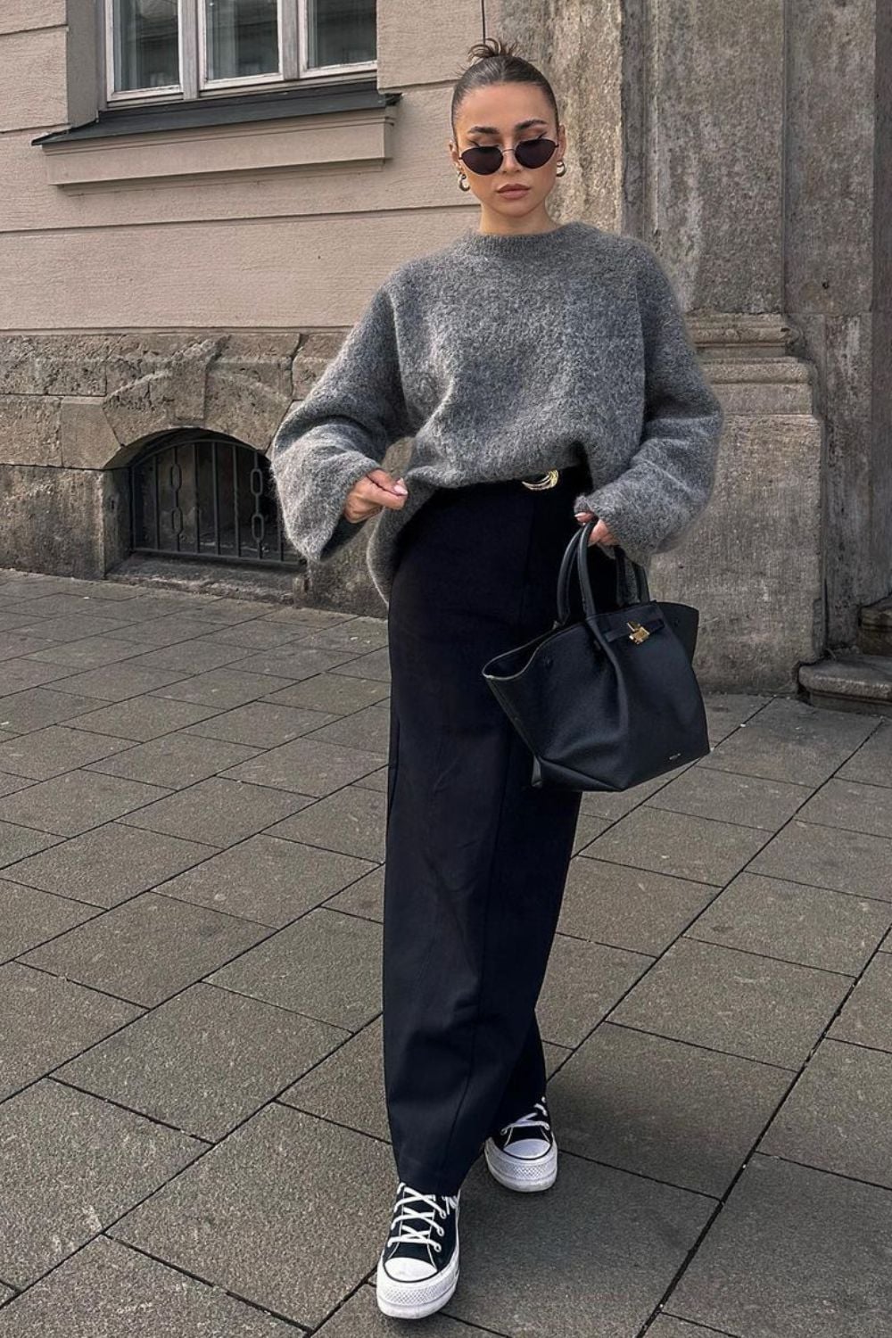 Woman wearing a loose gray sweater, black wide-leg pants, black sneakers, sunglasses, and gold hoop earrings, holding a black handbag while standing on a city sidewalk.