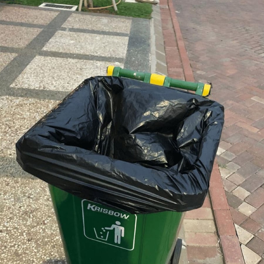 A green outdoor trash bin with a black plastic bag liner, placed on a sidewalk next to a paved street. The lid is open and rests against the back of the bin.