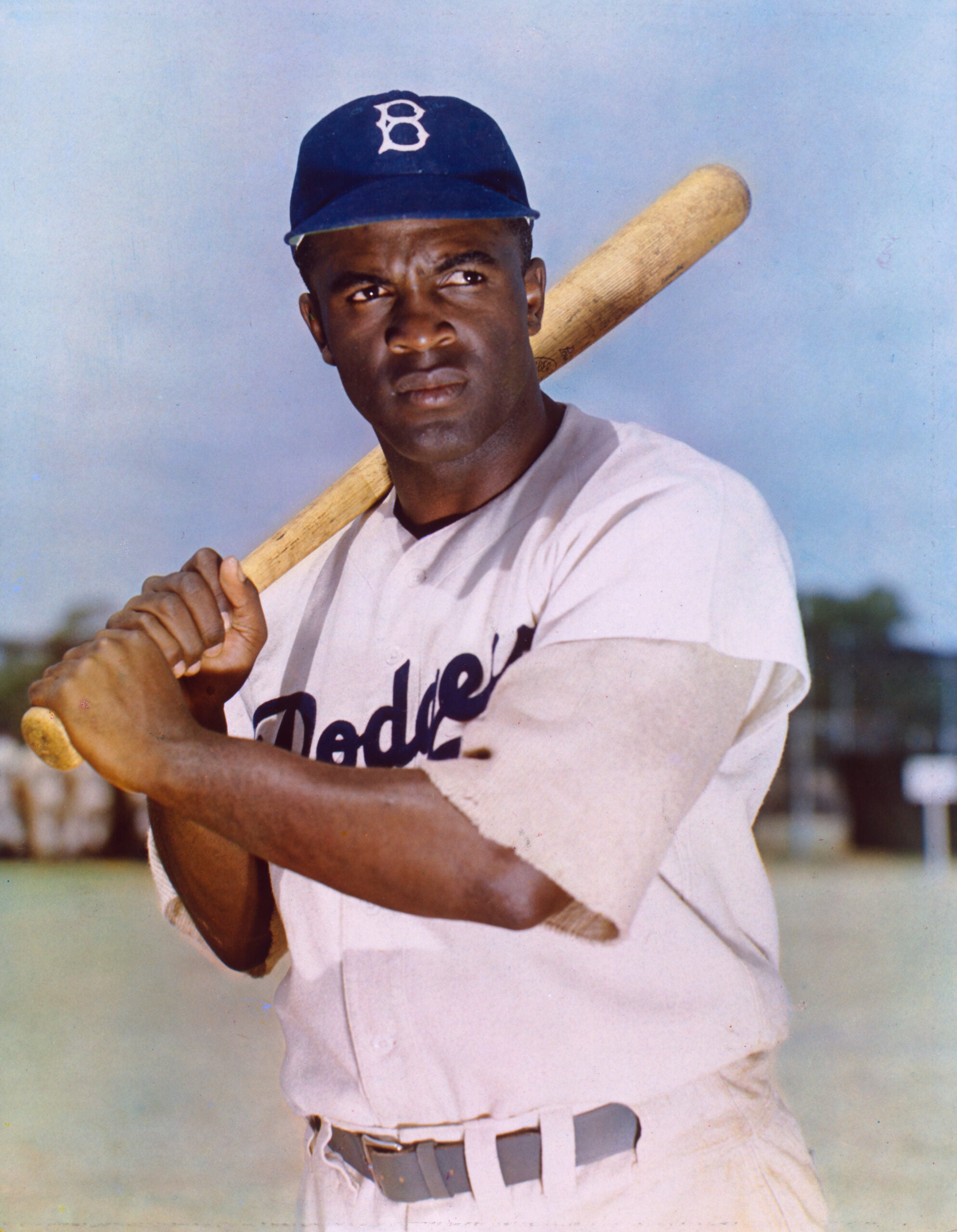 A baseball player in a Dodgers uniform holds a bat on his shoulder, standing outdoors on a field, looking into the distance.