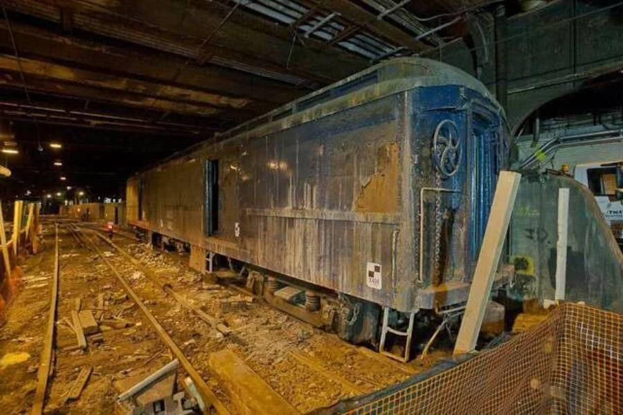 A weathered, graffiti-covered train car sits on rusty tracks in an underground, dimly lit tunnel surrounded by debris and construction materials.