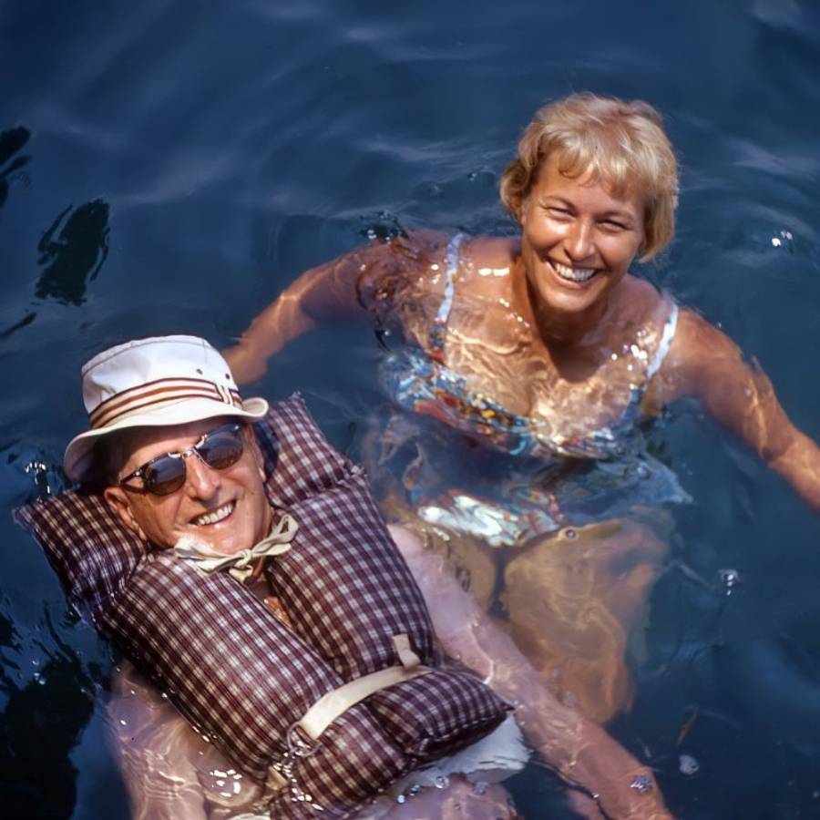 Two smiling adults are in the water. One person wears a hat, sunglasses, and a plaid life jacket, while the other is beside them in a floral swimsuit. Both appear to be enjoying a swim on a sunny day.