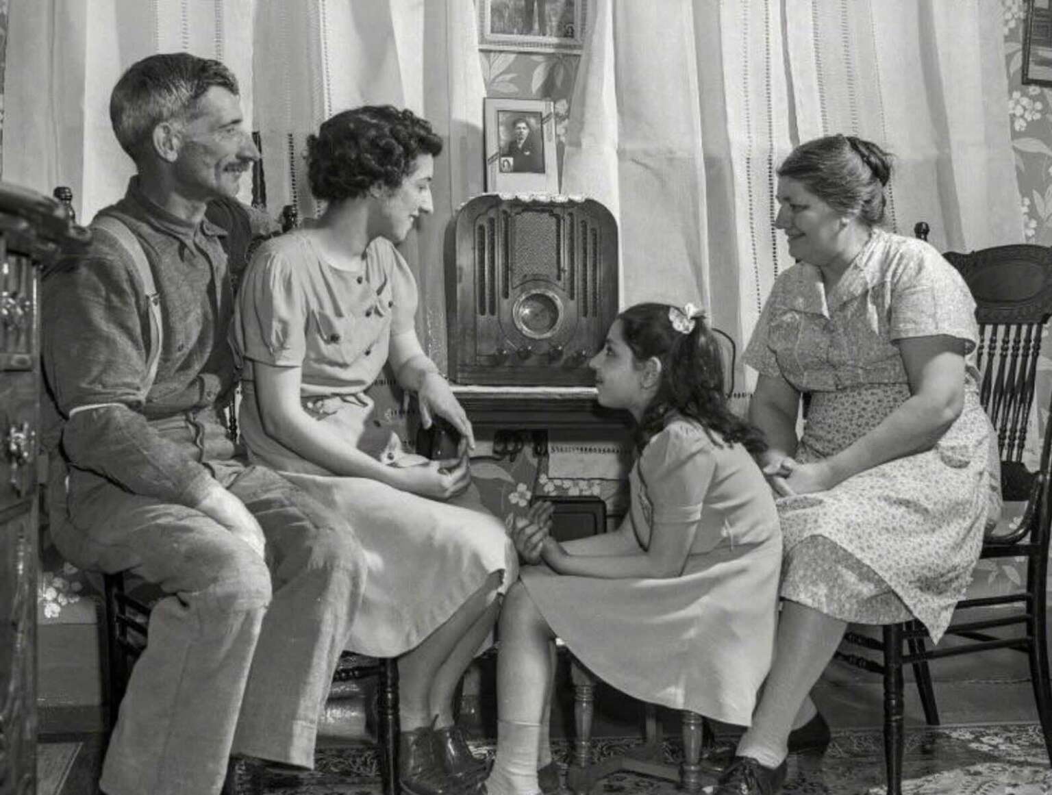 A vintage black-and-white photo shows a family of four, two adults and two girls, sitting around a radio in their living room, smiling and talking together. Sheer curtains and framed photos decorate the background.
