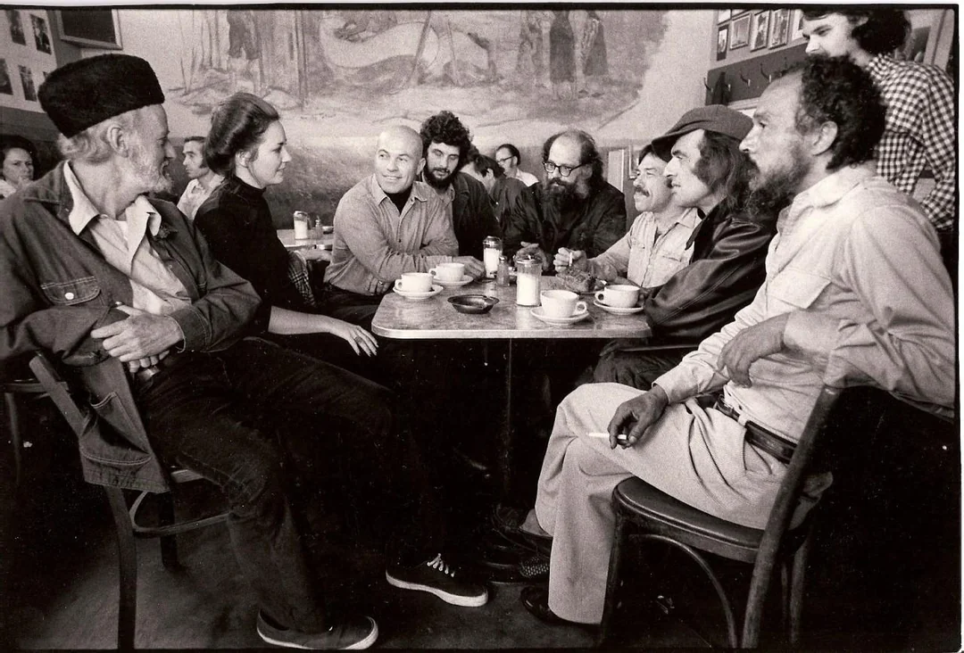 A group of people sit closely around a small café table, drinking coffee and conversing. The scene is lively, with an art-filled wall and framed photos in the background.