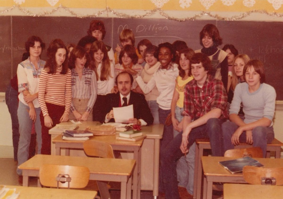 A group of students poses around a teacher seated at a desk in a classroom with a chalkboard and decorations, all smiling for a class photo.